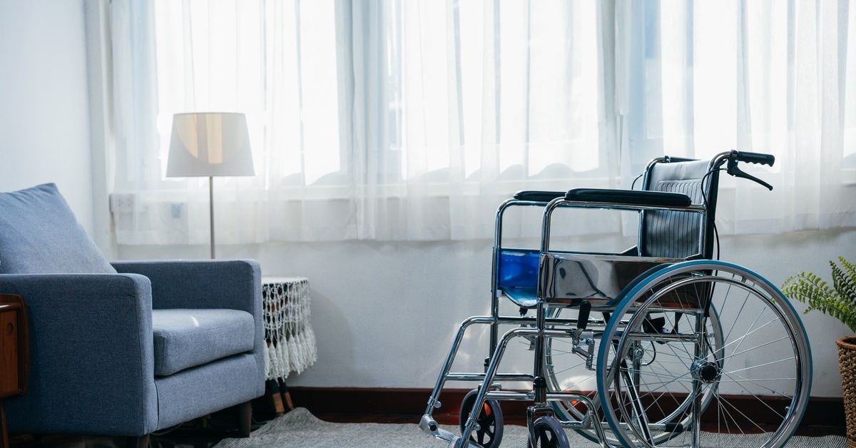 A room in a nursing facility with a large window and a sitting chair. In front of the chair is a wheelchair.