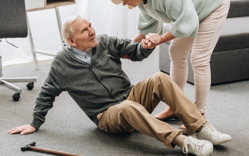 An elderly man lays on the ground receiving help from someone else to get back up. His cane sits next to him.
