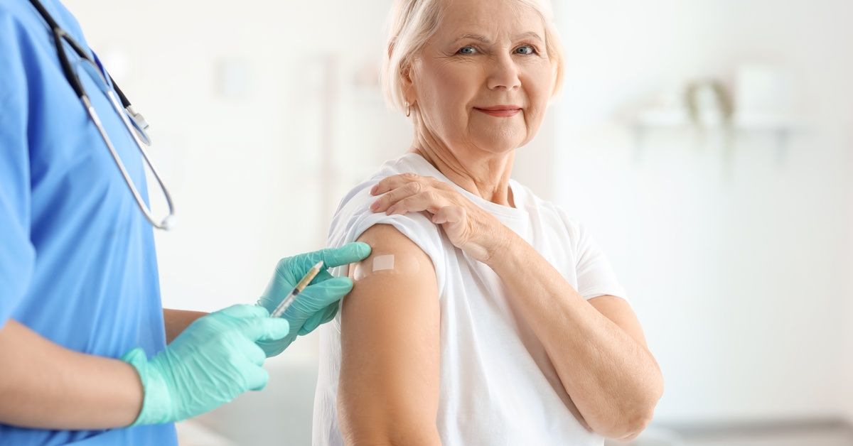 A senior woman sits on a medical table with a professional in blue scrubs standing next to her with a vaccine.