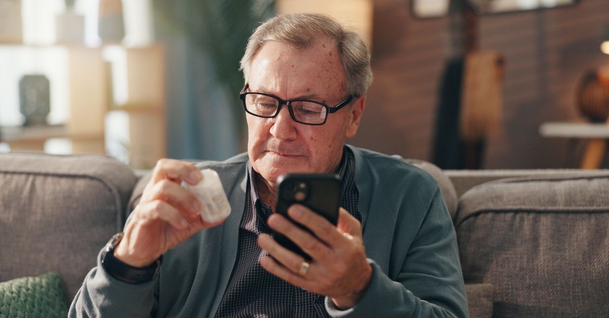 A senior man sits on the couch with his prescription bottle in one hand and his phone in the other, verifying information.