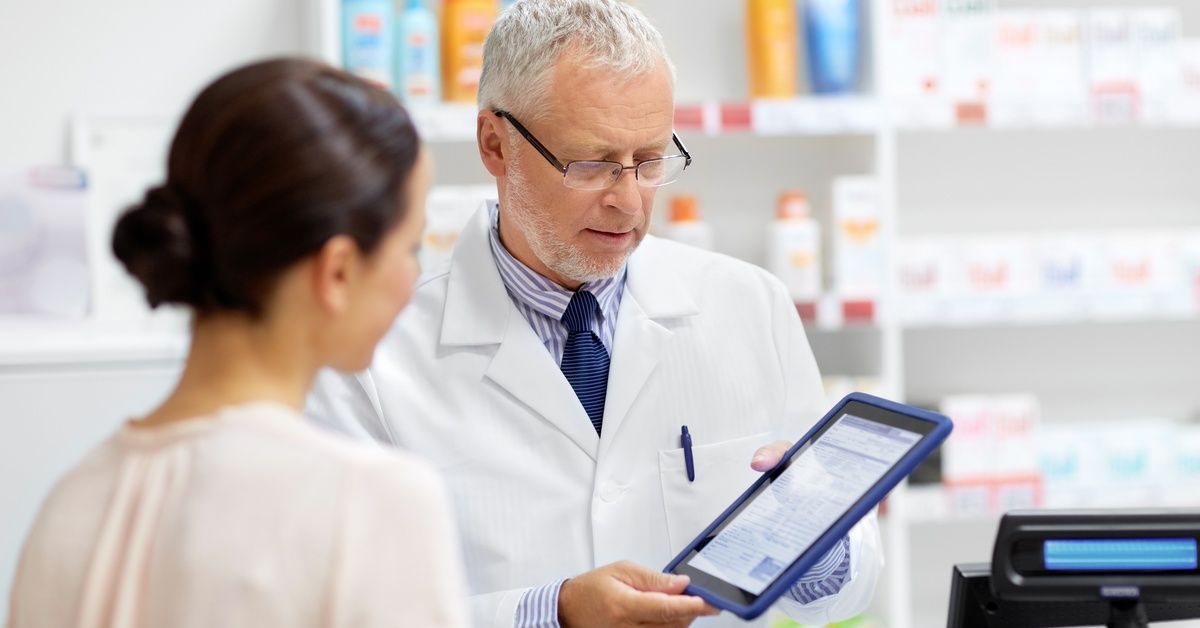 A male pharmacist holds a tablet in the pharmacy as he shares information with a pharmacy technician.