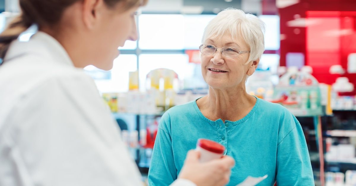 A senior pharmacy customer wears a teal blouse and discusses their medication with the pharmacist at the counter.