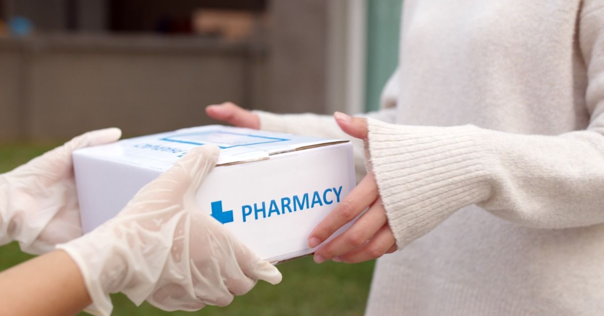 A close-up of a customer receiving a pharmacy delivery box from a person in a pair of white latex gloves.