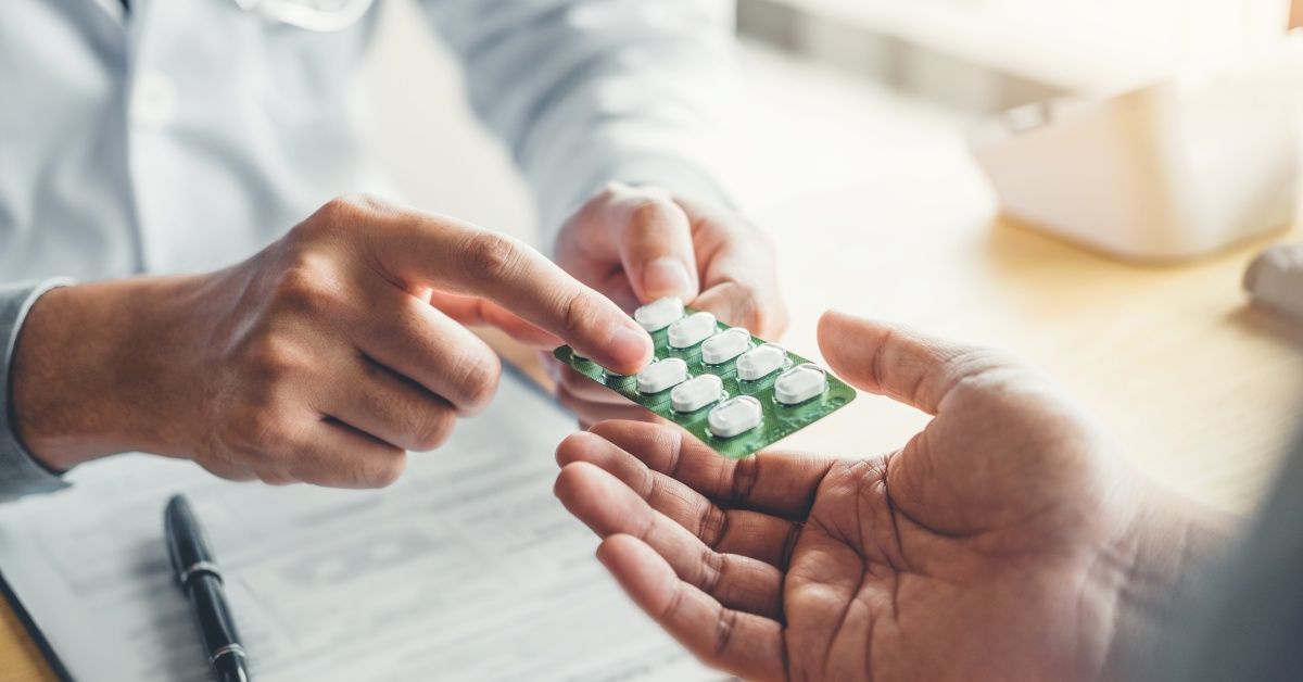 A pharmacist hands a blister pack of medication to a patient. The medication is white and oblong shaped.