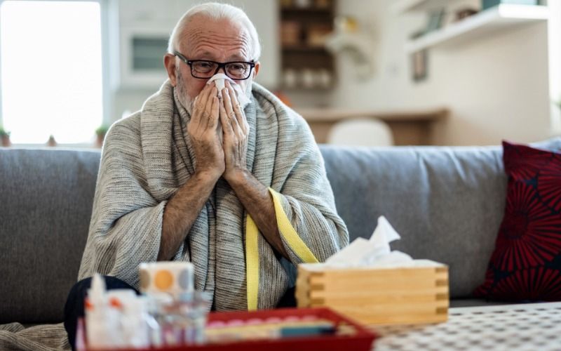 A senior man blows his nose into a tissue as he sits on the edge of his sofa in his home. He's wrapped in a blanket.
