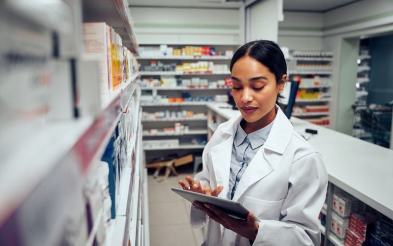 A young female pharmacist in her white lab coat uses a tablet to monitor inventory in the pharmacy aisle.