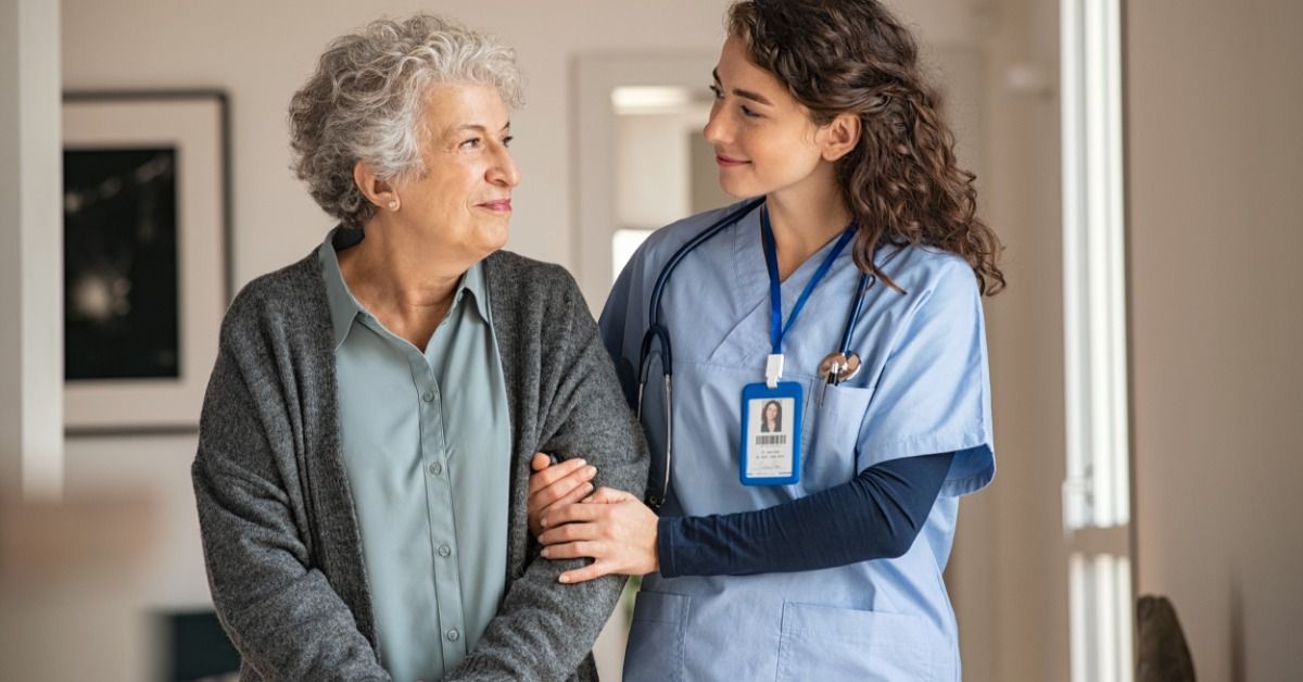 A long-term care facility nurse walks with a patient in the hallway. The nurse holds the patient's arm for support.