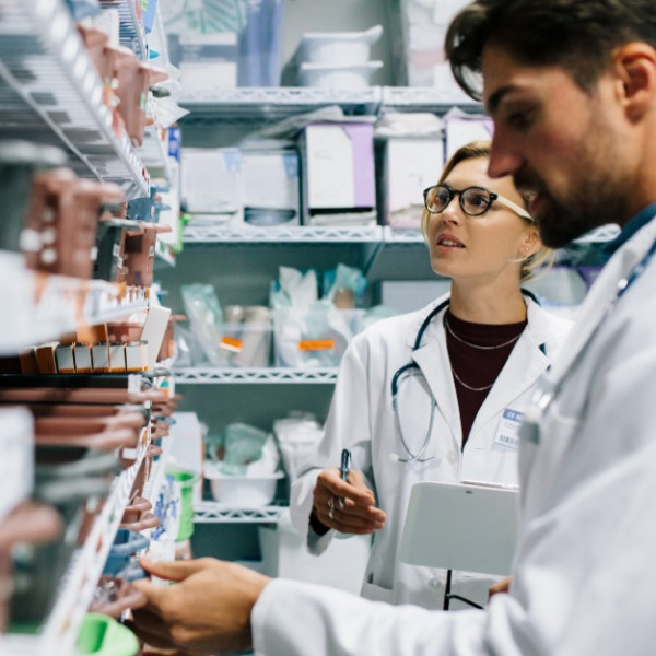Two pharmacy technicians stand in the aisle of a pharmacy as they discuss medication refills and orders.