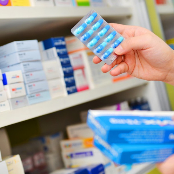 A close-up of a pharmacist in a medication aisle holding onto a blister pack of blue pills and a stack of boxes.