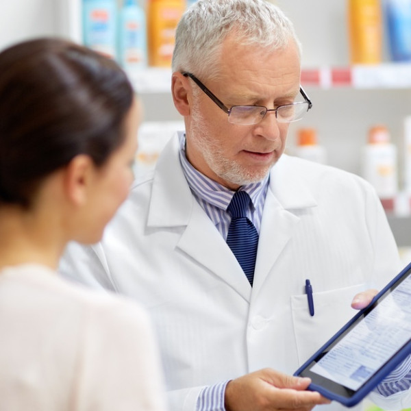 A male pharmacist holds a tablet in the pharmacy as he shares information with a pharmacy technician.