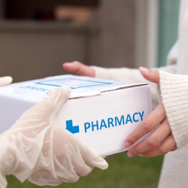 A close-up of a customer receiving a pharmacy delivery box from a person in a pair of white latex gloves.