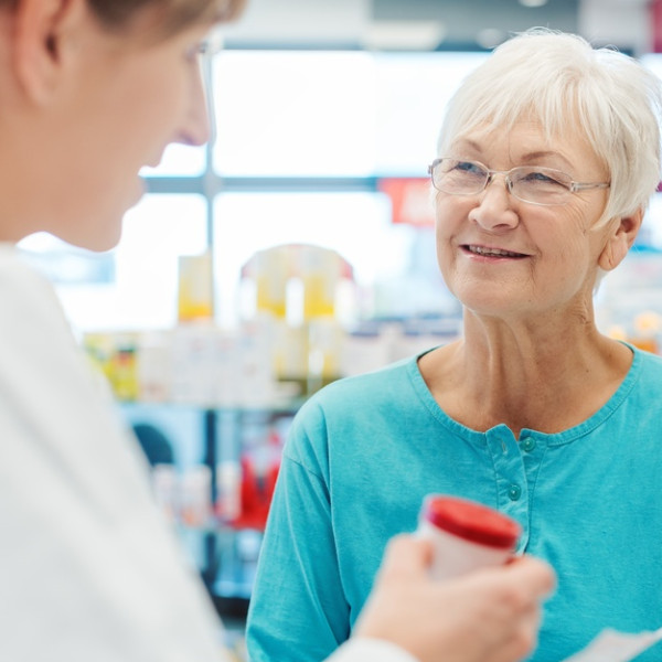 A senior pharmacy customer wears a teal blouse and discusses their medication with the pharmacist at the counter.