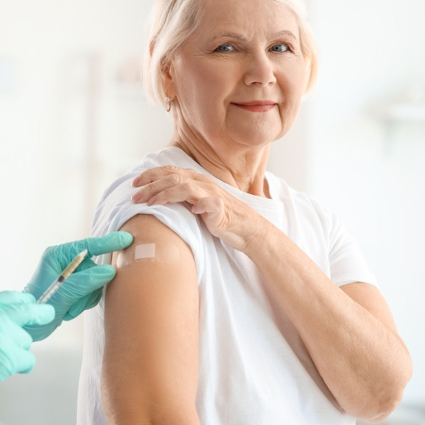 A senior woman sits on a medical table with a professional in blue scrubs standing next to her with a vaccine.