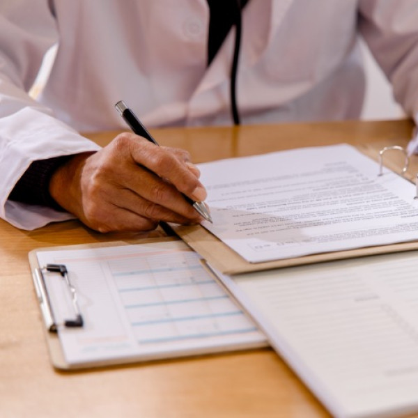 A medical professional sits at a desk to review a medication profile for a patient. They review a document on a clipboard.