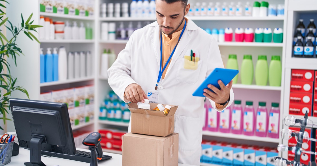 A male pharmacist stands at the register counter to pack a prescription delivery order. They pack two boxes.