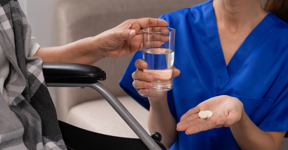 A close-up of a long-term care facility nurse handing medication and water to a patient in a wheelchair.