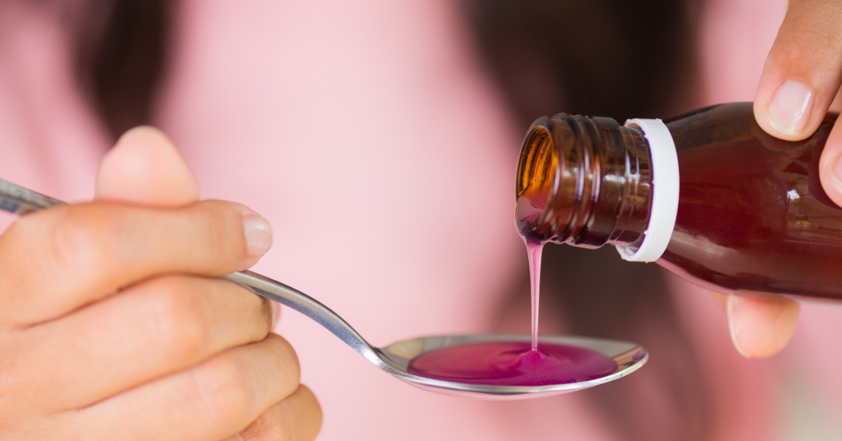 Someone pouring liquid medication from a brown prescription bottle onto a stainless steel kitchen spoon.