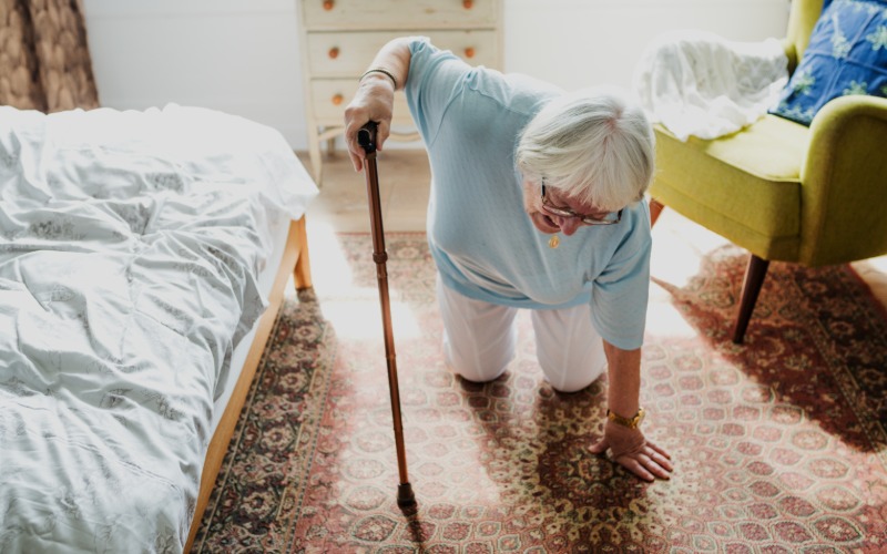 An elderly woman on her knees in her bedroom, struggling to get back up. She's using her cane for support.