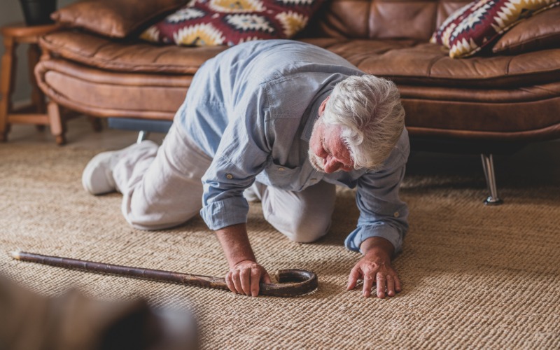 An elderly individual face down on the floor in front of their loveseat. They're using their cane to brace themselves.