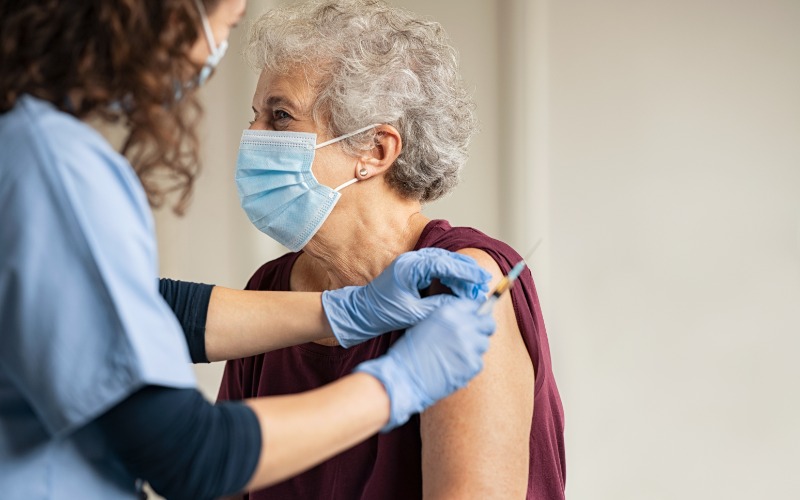 A senior woman sits close to a healthcare provider. The provider wears blue scrubs and administers a vaccine.