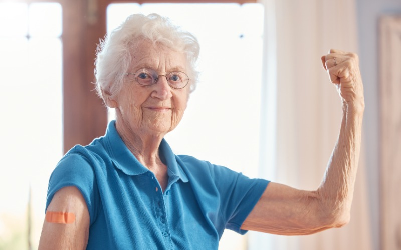 A senior woman in a blue polo shirt smirks as she flexes her right arm. Her left arm has an orange bandage from a vaccine.