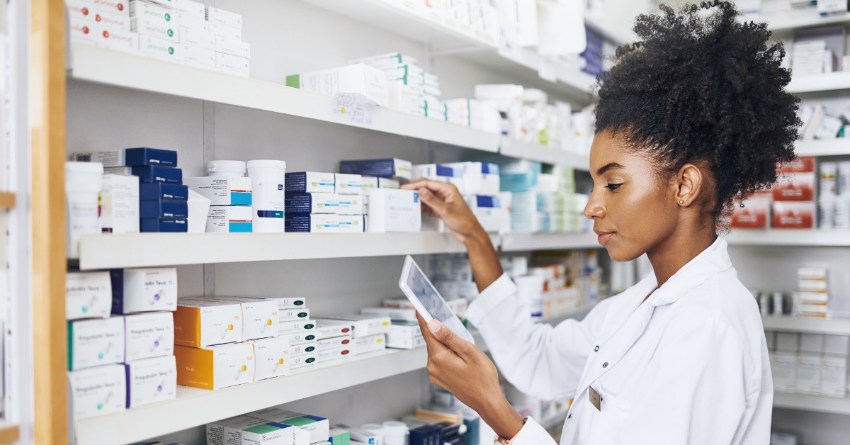 A young Black pharmacist stands at the inventory shelf to count boxes. She uses a tablet to reference the data.
