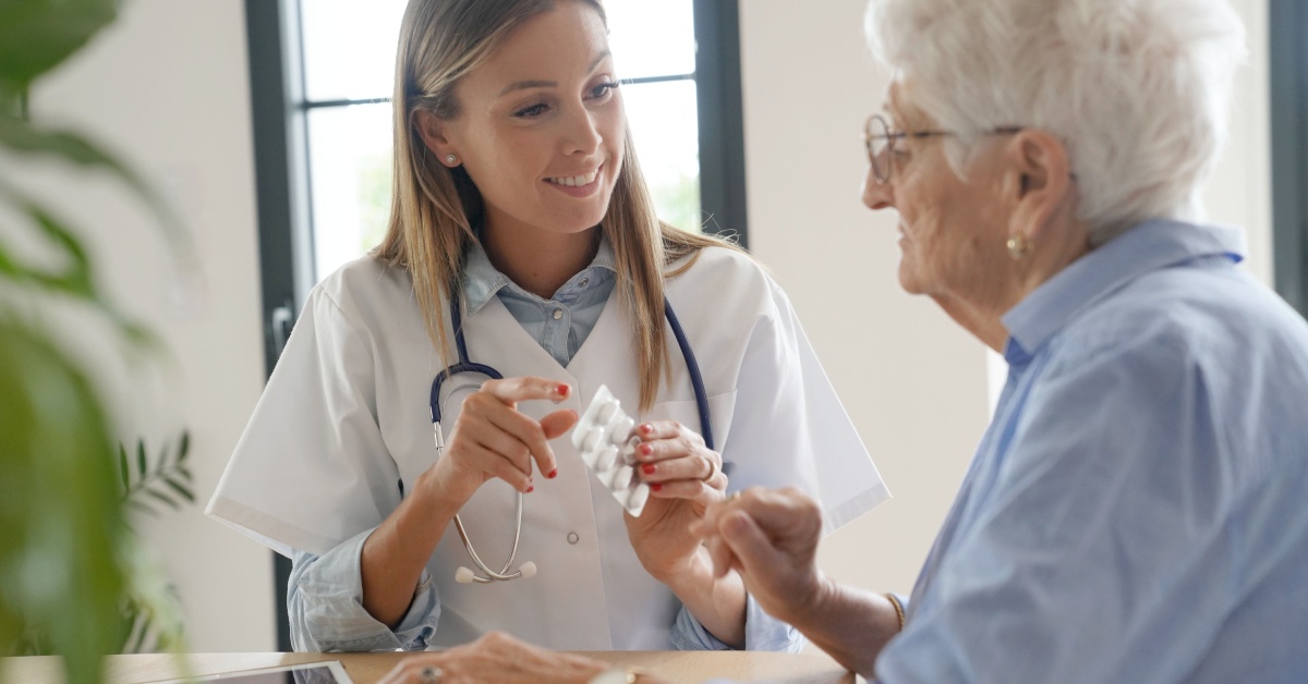 A young nurse hands a blister pack of medication to a long-term care facility patient. The nurse is smiling.