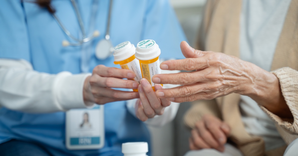 A nurse in blue scrubs sits next to an aging patient as the nurse hands over two pills bottles to the patient.