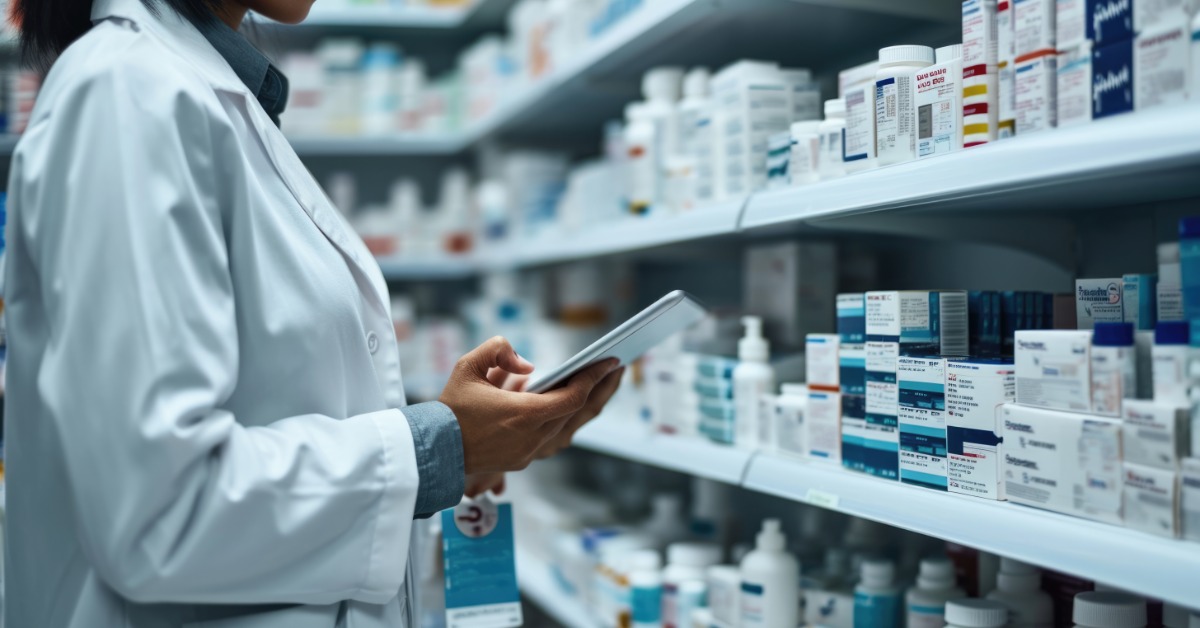 A pharmacist stands next to the medication inventory on the shelves with a tablet to confirm inventory counts.