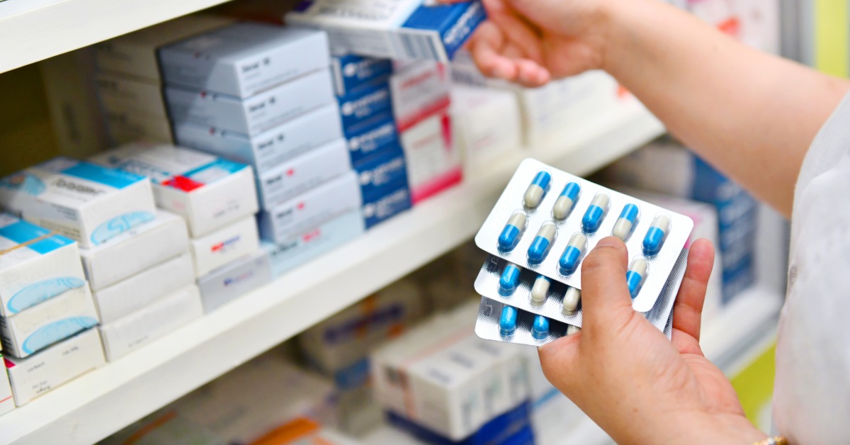 A close-up of a pharmacy technician holding a foil packet of medication in their left hand. They also grab a med box.