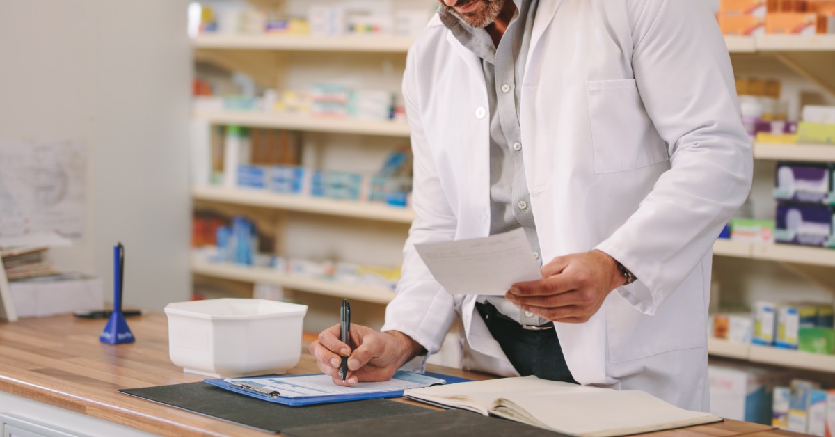 A male pharmacist stands at the pharmacy counter in his lab coat. He takes notes on a notepad and reads a paper.