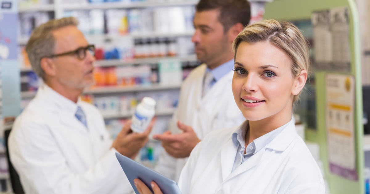 Three pharmacists stand in the pharmacy to discuss a white medication bottle. One pharmacist is smiling.