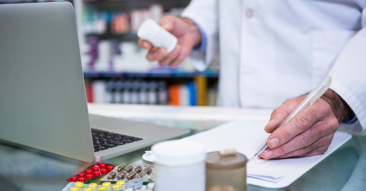 A pharmacist in a white coat works at the counter with paperwork, medication bottles, and a laptop. They're writing.