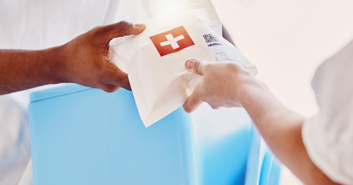 A person hands over a prescription delivery bag with a red cross on the packaging. Another person grabs the package.