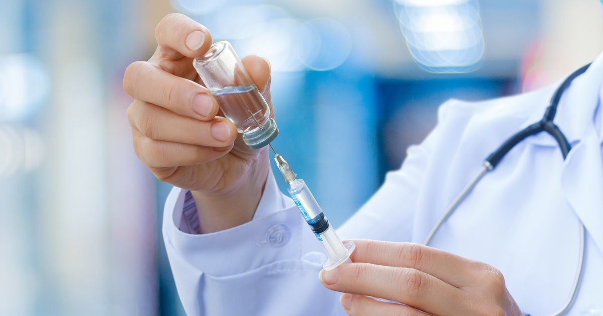 A close-up of a medical provider in a white coat and stethoscope holding a vaccine bottle and syringe.