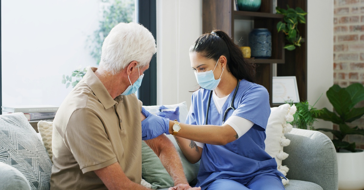 A masked senior sits with a masked provider in blue scrubs as the provider administers a vaccine to the patient.