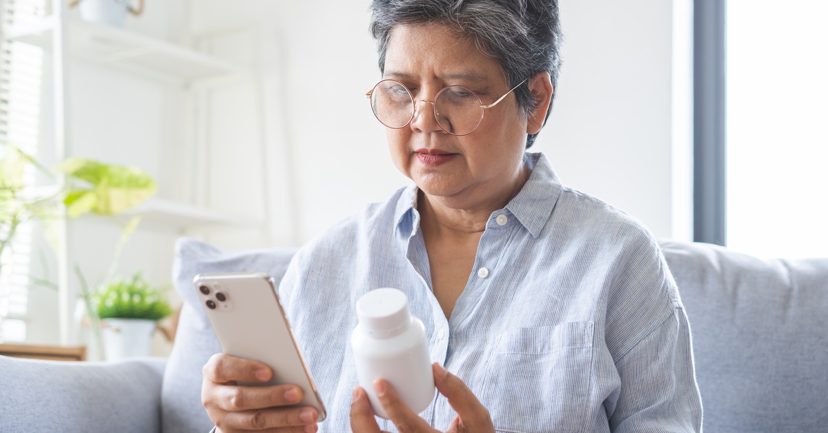 A senior woman sits on the couch at home holding a medicine bottle in one hand and her phone in the other.