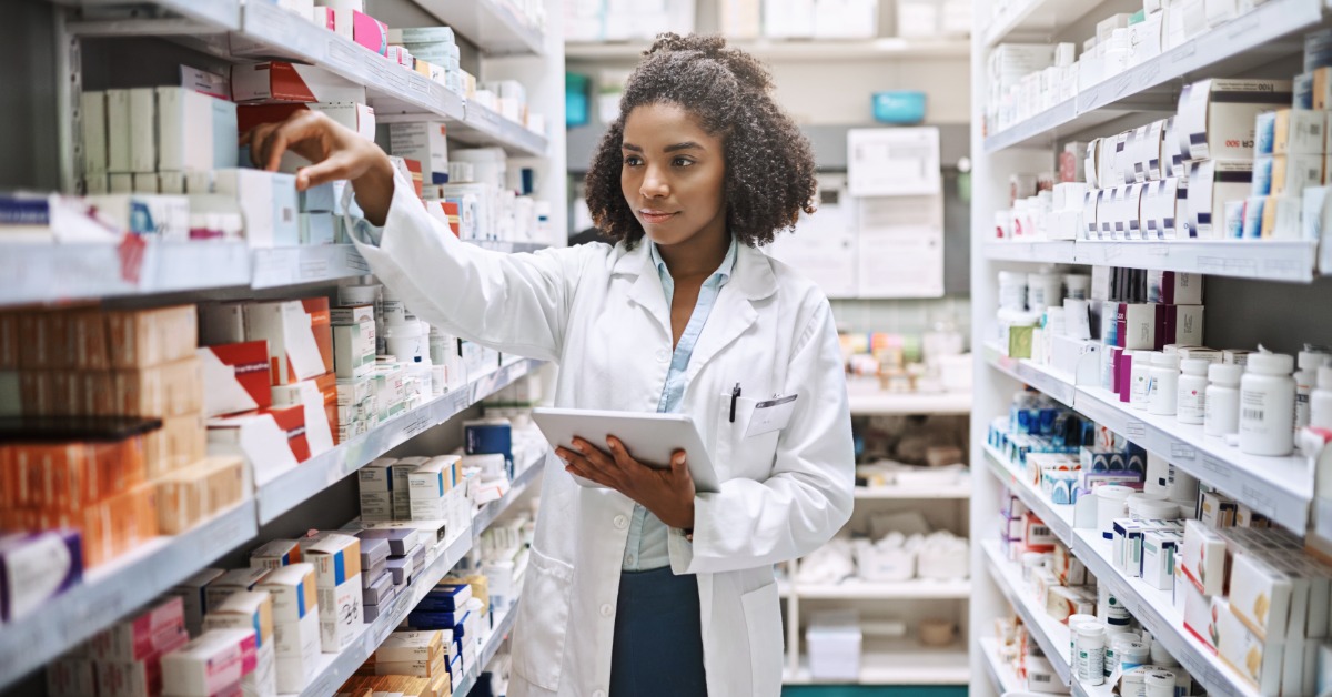 A young Black woman works at the pharmacy wearing a white coat and holds a tablet to check inventory on the shelf.