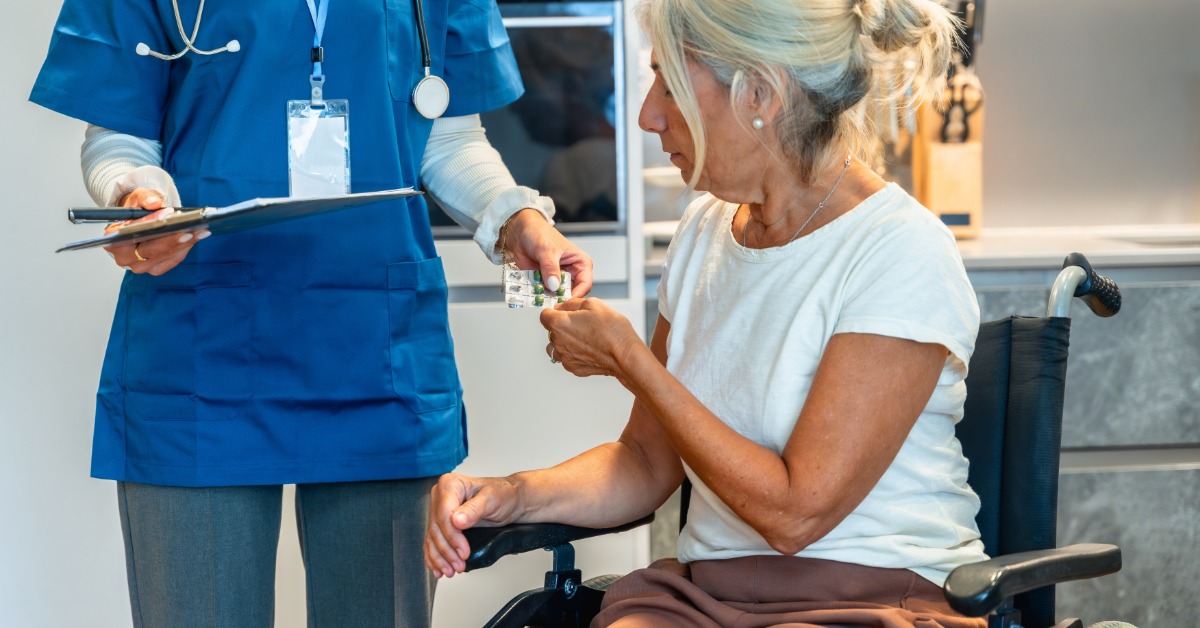A nurse in a blue scrub top holds a clipboard and hands a senior woman in a wheelchair a blister pack of medication.