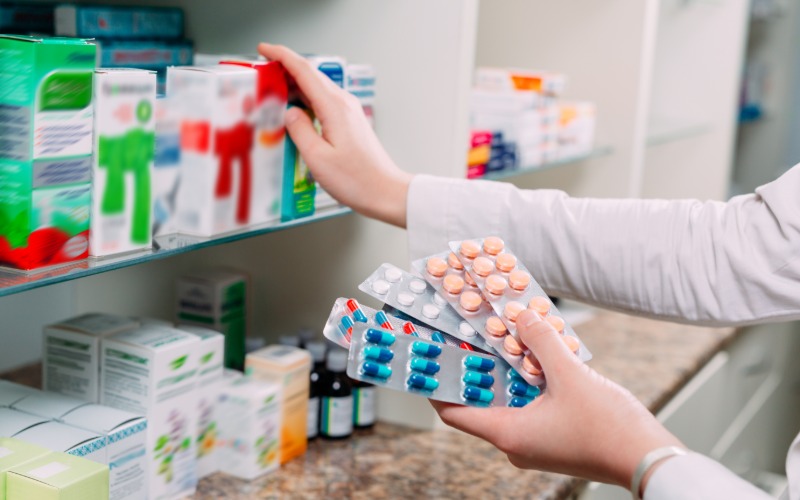 A pharmacist pulls inventory off the shelf as they hold several blister packs in the other hand. The pills are multi-color.