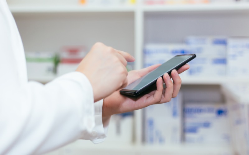 A close-up of a pharmacist holding a smartphone as they check the mobile Rx app for prescription updates.