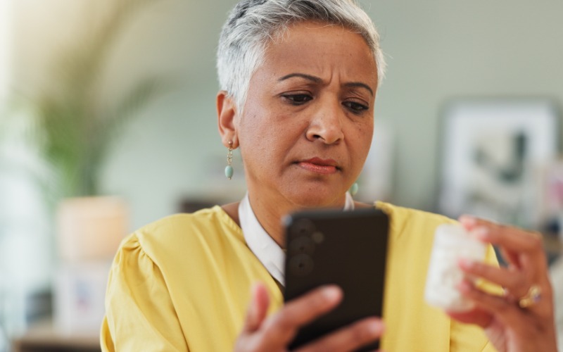 A senior woman in a yellow top holds her phone as she reads her medication bottle. She's using the mobile delivery app.