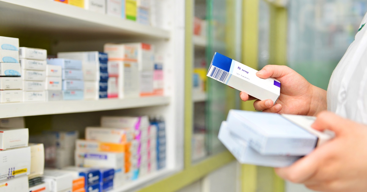 A pharmacist's hands near the shelf holds a few medication boxes. There are different meds on the shelf.