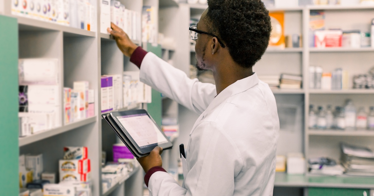 A Black pharmacist stands at the inventory shelf with a tablet and his white coat on to count stock levels.