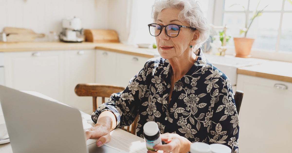 A senior woman sits at her dining room table to order new medication on her laptop. She wears glasses.